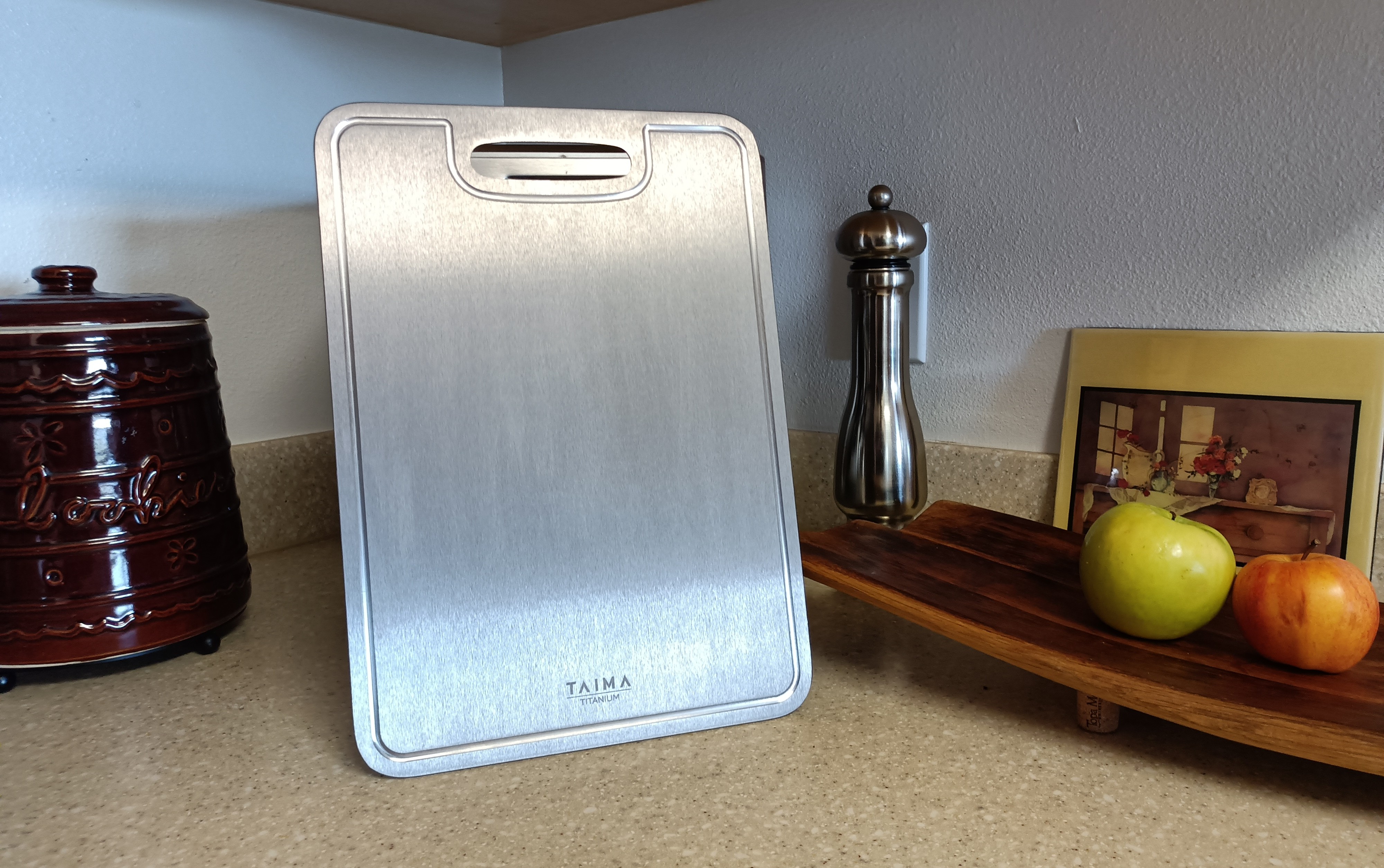 Taima Titanium Cutting Board V2 in a real kitchen setting, standing upright on a counter beside produce and everyday tools