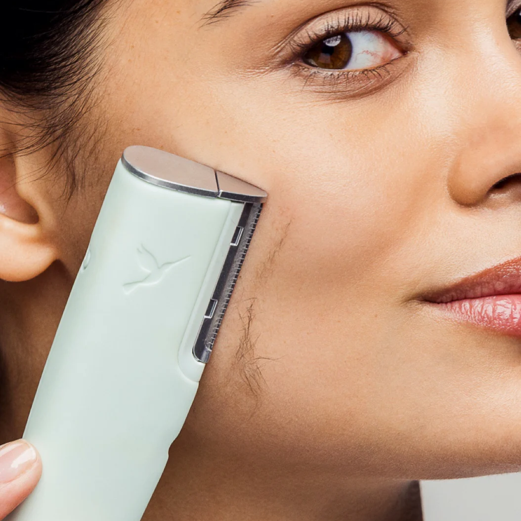 Woman gently gliding a dermaplaning device along her cheek to exfoliate dead skin and remove peach fuzz